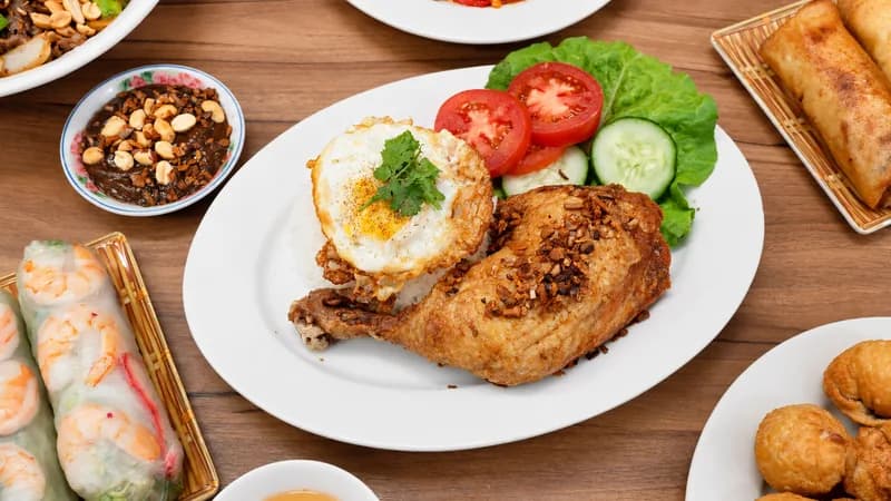 Grilled chicken rice platter with a fried egg, surrounded by fresh spring rolls, peanut sauce, egg rolls and fried wontons on a wood table